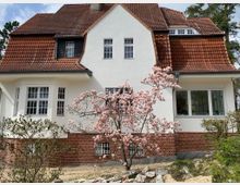 Ein zweigeschossiges Haus im traditionellen Stil mit weißer Fassade, roten Ziegeldächern und Sprossenfenstern. Im Vordergrund steht ein blühender Baum mit rosa Blüten, umgeben von einem Gartenbereich mit Sträuchern.
