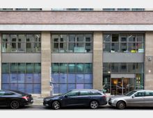 Fassadenansicht eines modernen Bürogebäudes mit großen Glasfenstern und einem Eingangsbereich, flankiert von geparkten Autos entlang der Straße. Die oberen Etagen und der Backsteinabschluss kontrastieren mit der Betonstruktur der unteren Stockwerke.