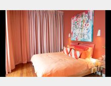 Schlafzimmer mit orangenen Wänden und einem Holzboden, ausgestattet mit einem Doppelbett in orangefarbenen Textilien und dekorativen Kissen. Im Hintergrund hängen lange, rosafarbene Vorhänge sowie ein farbenfrohes Kunstwerk mit Schriftzug.