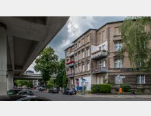 Straßenszene in einem städtischen Wohngebiet mit einem historischen Mehrfamilienhaus aus Stein auf der rechten Seite, dekoriert mit Blumen auf Balkonen. Links im Bild verläuft eine erhöhte Schnellstraße, darunter parkende Autos, sowie Bäume und eine Gehwegführung für Fußgänger.