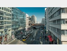 Straßenszene in einer Stadt mit modernen Gebäuden auf beiden Seiten, einer gelben Straßenbahn in der Mitte und einer erhöhten S-Bahn-Station im Hintergrund (Bahnhof Friedrichstraße). Zahlreiche Fußgänger beleben die Gehwege entlang der Geschäfte und Cafés.