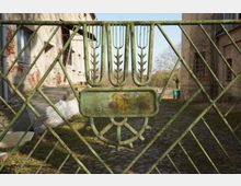 Nahaufnahme eines verwitterten, grün lackierten Metalltors mit rautenförmigem Gittermuster und dekorativem Emblem in der Mitte. Dahinter liegt ein gepflasterter Hof zwischen alten, teilweise beschädigten Gebäudefassaden.