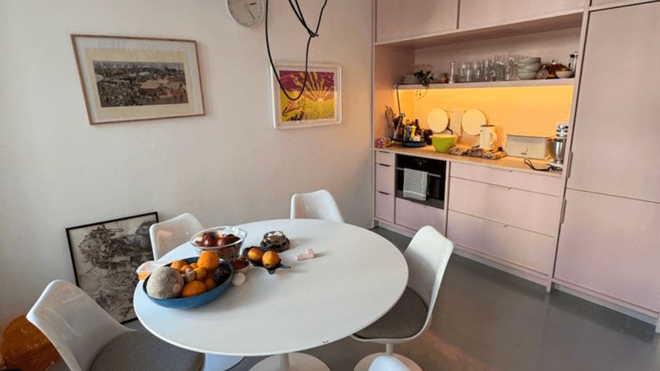 Modern open-plan kitchen and dining area with pale pink built-in cabinets and a lit backsplash along one wall. A round white dining table with white pedestal chairs sits under a large hanging pendant light, with framed artwork on the white walls and a gray floor.