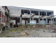 Verfallenes Industriegebäude mit bröckelnden Ziegel- und Betonwänden, teils zerstörten Fenstern und Graffiti auf den Fassaden. Der verwilderte Vorplatz ist mit Moos und Unkraut bewachsen.