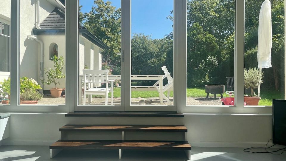 Blick aus einem hellen Wohnraum durch große, bodentiefe Fensterfront auf eine Terrasse mit weißen Gartenmöbeln, Pflanzkübeln und einem runden Feuerkorb. Dahinter liegt ein grüner Garten mit dichtem Baumbestand; innen steht vor den Fenstern ein niedriger Holzstufen-Sockel.