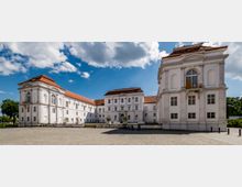 Helles Barockgebäude mit weißen Fassaden, roten Ziegeldächern und zahlreichen großen Fenstern, das einen rechteckigen Innenhof mit gepflastertem Boden umschließt. Der Himmel ist blau mit vereinzelten Wolken, und im Hintergrund befinden sich einige Bäume.