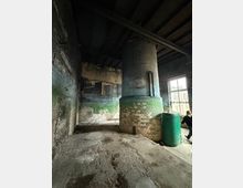 Innenraum eines verlassenen Industriegebäudes mit rauen, teils abgeplatzten Ziegel- und Putzwänden und einer dunklen Holzbalkendecke. In der Mitte steht ein großer zylindrischer gemauerter Ofen oder Schornstein, daneben ein grüner Tank; rechts fällt Licht durch ein großes Fenster.
