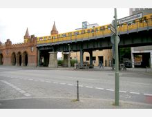 Backsteinbrücke mit gotischen Bögen und Ziegeltürmen, auf der eine gelbe U-Bahn auf einem erhöhten Schienennetz fährt; Straßenszene mit Verkehrsschildern und Gehweg im Vordergrund, umgeben von modernen Gebäuden.