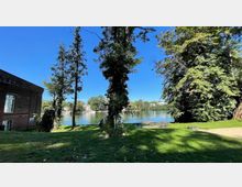 Grüne Parklandschaft mit Blick auf einen ruhigen Fluss, flankiert von Bäumen und einem Backsteingebäude auf der linken Seite. Im Hintergrund sind weitere Gebäude am gegenüberliegenden Ufer sichtbar.