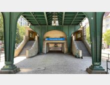 Eingangsbereich der U-Bahnstation "Eberswalder Straße" in Berlin unter einer grünen Stahlkonstruktion einer Hochbahn, mit beidseitigen Treppenaufgängen und einem blauen Stationsschild über dem Durchgang. Im Hintergrund sind Bäume und umliegende Gebäude sichtbar.