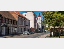 Straßenszene einer deutschen Kleinstadt mit historischem Fachwerkhaus, mehrstöckigen Gebäuden mit roten Ziegeldächern und einem weißen Turmgebäude mit spitzer Dachform im Hintergrund; rechts im Bild eine Baumreihe und eine Bushaltestelle.