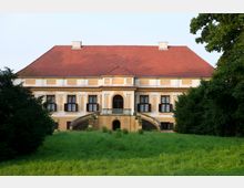 Historisches Gebäude mit einem roten Walmdach und einer symmetrischen Fassade, die mit Stuckelementen verziert ist. Vor dem Eingang befinden sich zwei halbkreisförmige Treppen, die in eine von Bäumen umgebene Rasenfläche führen.