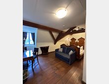 Ein gemütliches Zimmer mit Holzbalkendecke, Holzboden und antiken Möbeln, darunter ein Doppelbett mit verzierten Kopfteilen, ein blauer Zweisitzer und ein Holztisch mit Stühlen. Ein Fenster mit Vorhängen lässt Tageslicht herein.