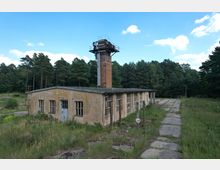 Ehemaliges Industriegebäude mit verfallener Fassade und zerbrochenen Fenstern, neben einem hohen Backsteinturm mit Metallplattformen, umgeben von dichter Waldlandschaft und verwildertem Gras. Ein alter, betonierter Weg führt am Gebäude vorbei.