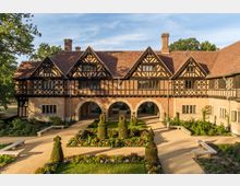 Historisches Gebäude im Tudor-Stil mit aufwändiger Fachwerkfassade, rotem Ziegeldach und drei großen Steinbögen in der Mitte. Im Vordergrund befindet sich ein symmetrisch gestalteter Garten mit geschnittenen Buchsbaumfiguren, Blumenbeeten und Sitzbänken.