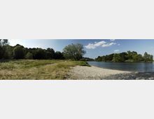 Flussufer mit sandigem Strand, umgeben von Wiesen und dichter Vegetation aus Bäumen und Sträuchern; im Hintergrund befindet sich ein ruhiger Fluss unter einem blauen Himmel mit wenigen Wolken.