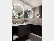 Modernes Badezimmer mit grauen Wänden und großflächigen Spiegeln, die den Raum reflektieren. Ein Waschtisch mit dunklem Unterschrank, weißer Ablage, eingelassener Armatur, sowie leuchtende, runde Spiegel prägen das Design.