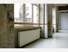 Ein heller Tanzraum mit großen Fenstern, weißen Vorhängen und einer Ballettstange, die an einer freiliegenden Backsteinmauer befestigt ist. Ein Heizkörper befindet sich unter den Fenstern, und der Boden ist aus hellem Material.