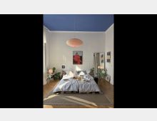 Ein helles Schlafzimmer mit hohen weißen Wänden und einer blauen Decke. Im Mittelpunkt steht ein niedriges Doppelbett mit grauer Bettwäsche, umgeben von Pflanzen, einem Teppich und kleinen Nachttischen; an der Wand hängen dekorative Bilder.