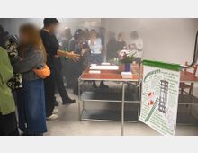 Eine Gruppe von Menschen steht in einem hell beleuchteten Raum mit weißen Wänden, während im Vordergrund ein Tisch mit Schriftmaterialien und einem Blumenarrangement aufgestellt ist. Rechts hängt ein Plakat mit Veranstaltungsinformationen an einem Metallständer.