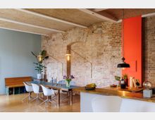 Wohnzimmer mit Ess- und Küchenbereich in industriellem Stil. Sichtbare Ziegelwand, Holzfußboden, eine lange Holztischgruppe mit weißen Stühlen und eine offene Küchenzeile mit Hängelampe; orangefarbene Wandakzente und dekorative Pflanzen.