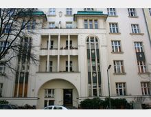 Wohngebäude im Stil des frühen 20. Jahrhunderts mit cremefarbener Fassade, dekorativen Ornamenten und hohen Fenstern. Das Gebäude verfügt über Balkone mit Blumenkästen, einen bogenförmigen Eingangsbereich und ein grünes Dach sowie symmetrisch angeordnete Fenster und vertikale Zierelemente.