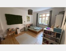 Ein Schlafzimmer mit Holzboden, einem einfachen Holzrahmenbett mit Bettwäsche, einem großen Fenster mit Vorhängen und einer weißen Heizung darunter. An der Wand hängt ein künstlicher Pflanzendekor sowie ein kleines Poster, und ein Regal auf der rechten Seite enthält Bücher und Dekorationsgegenstände.