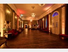 Eleganter langer Saal mit Holzfußboden, Kronleuchtern und Wandleuchtern, dekoriert mit roten Samtvorhängen und antiken Möbeln. Die gold-beigen Wände und Dekorationen schaffen eine warme, atmosphärische Beleuchtung.