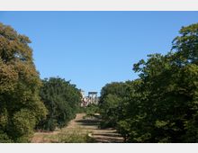 Weitläufiger Park mit einer offenen Rasenfläche, gesäumt von großen Bäumen; im Hintergrund ist eine historische Ruine mit Säulen und Fragmenten antiker Architektur zu sehen, vor einem klaren blauen Himmel.