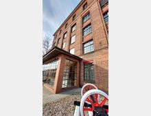 Backsteinbau mit großen Fenstern und rotem Fensterrahmen, ergänzt durch einen gläsernen Eingangsbereich. Im Vordergrund sind ein Kiesbereich und ein rot-weißes Maschinenteil zu sehen.