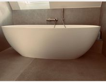 Freistehende weiße Badewanne in einem Badezimmer mit grauen Wandfliesen und beige Bodenfliesen. Im Hintergrund befindet sich ein kleines Fenster unter einer schrägen Decke.