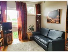 Wohnzimmer mit einer schwarzen Ledercouch, roten Vorhängen mit kreisförmigem Muster, einem kleinen Holztisch mit Pflanze, und einem weißen Teppich auf Holzboden. An der Wand hängt ein großes Bild, daneben steht ein TV auf einem Holzschrank.