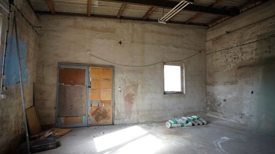 Innenraum einer unfertigen Werkstatt oder Garage mit rohen Beton- und verputzten Wänden, freiliegender Decke mit Holzbalken und einer hängenden Leuchtstofflampe. An der Rückwand stehen eine provisorisch verkleidete Tür und ein kleines Fenster; auf dem Betonboden liegen einige Bausäcke und Baumaterialien.