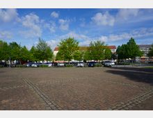 Gepflasterter Parkplatz mit vereinzelten, begrünten Baumreihen im Vordergrund; im Hintergrund stehen mehrstöckige Wohngebäude mit roten Dächern und ein Gebäude mit Solarpanelen. Der Himmel ist blau und teils wolkig.