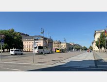 Straßenszene in einer historischen Innenstadt mit breitem Gehweg, mehreren Gebäuden im klassizistischen Stil und einer gepflasterten Straße. Im Vordergrund geparkte Busse und Autos, im Hintergrund Bäume und Laternen im historischen Design.
