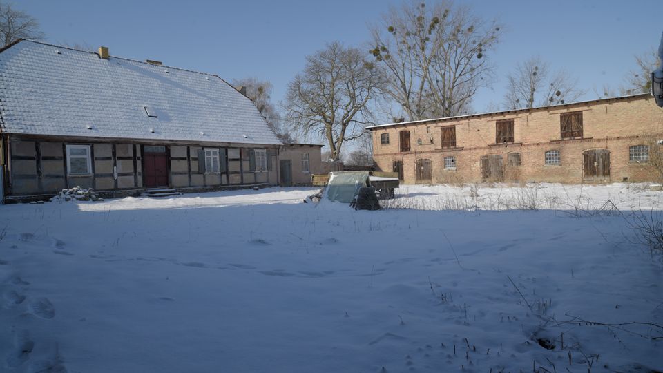 Verschneiter Bauernhofhofplatz mit einem Fachwerkhaus mit schneebedecktem Satteldach links und einem langen Backstein-Stallgebäude mit Holztoren rechts. Im Vordergrund liegt eine weite Schneefläche mit wenigen Grashalmen, dahinter stehen kahle Bäume vor blauem Himmel.