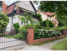 Wohnstraße mit zwei nebeneinander stehenden, stilvollen Einfamilienhäusern. Vor den Gebäuden befinden sich ein Vorgarten mit Blumen und Sträuchern, ein schmiedeeiserner Zaun sowie ein Weg mit Kopfsteinpflaster.