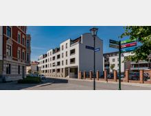 Straßenszene mit einem modernen Wohngebäude aus hellem Putz, flankiert von einem historischen Gebäude aus rotem Backstein und weißen Stuckelementen. Im Vordergrund befinden sich Straßenschilder, eine Laterne und ein gepflasterter Gehweg.