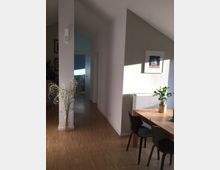 Helles Wohnzimmer mit Holzboden, einem Esstisch aus Holz mit schwarzen Stühlen und dekorativen Pflanzen. Im Hintergrund ein Flur mit weißen Wänden und einer geöffneten Tür, die zu einem blauen Raum führt.