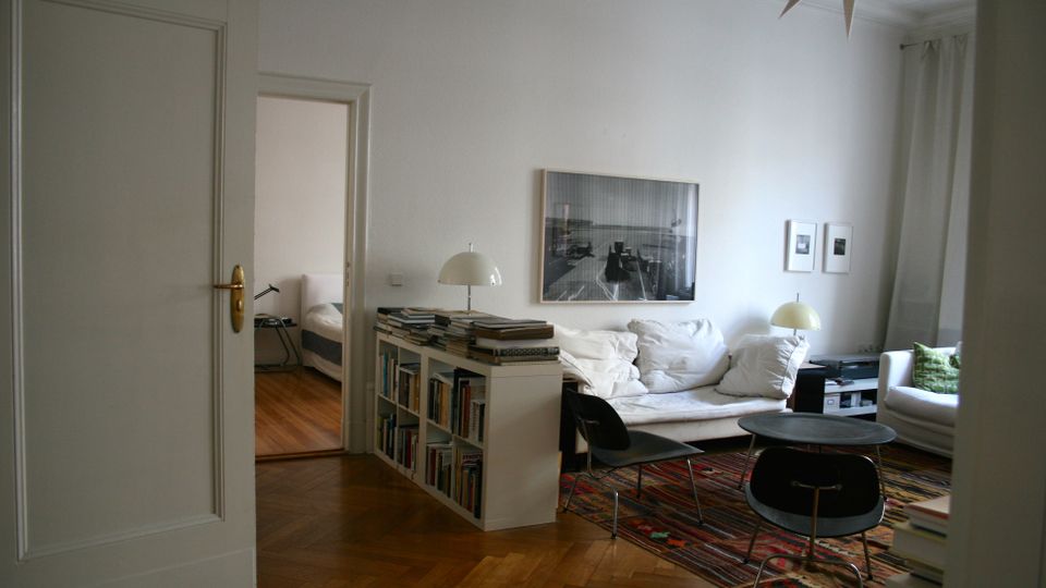 Heller Wohnraum mit Parkettboden, weißem Sofa, Couchtisch und buntem Teppich, dazu ein Bücherregal als Raumteiler und mehrere Tischlampen. An der Wand hängen gerahmte Fotos, und durch eine offene Tür ist ein angrenzendes Schlafzimmer zu sehen.