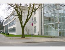 Modernes Bürogebäude mit großen Fenstern und einem Treppenhaus aus Glas an der Ecke von zwei Straßen, gekennzeichnet durch Straßenschilder "Gimritzer Weg" und "Aroser Allee". Im Vordergrund stehen ein Gehweg, ein Baum und ein Rasenbereich.