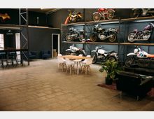 Ein großer Raum mit dunklen Wänden, in dem mehrere Motorräder auf einem Metallregal gestapelt sind. Im Vordergrund steht ein Esstisch mit weißen Stühlen, daneben befindet sich eine Sitzecke mit Sofas und einem Teppich, sowie Pflanzen als Dekoration.