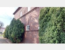 Backsteinfassade eines Gebäudes mit kleinen Fenstern, flankiert von hohen, dicht gewachsenen Koniferen. Im Vordergrund ist ein gepflasterter Weg zu sehen.