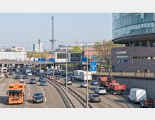Mehrspurige Autobahn in einem städtischen Gebiet mit hohem Verkehrsaufkommen, umgeben von moderner und industrieller Architektur. Im Hintergrund sind der Berliner Funkturm sowie mehrere Hochhäuser zu sehen, während Schilder für Ziele wie Magdeburg, Leipzig und die Messe Berlin ICC den Weg weisen.