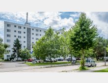 Mehrstöckiges Wohngebäude mit weißer Fassade und rechteckigen Fenstern, umgeben von Bäumen und Grünflächen. Im Vordergrund sind eine Parkfläche mit parkenden Autos und ein Bürgersteig zu sehen.