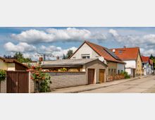 Reihen von Einfamilienhäusern mit roten Ziegeldächern in einer ruhigen Wohnstraße; einige Häuser haben Solarpanels auf den Dächern, Vorgärten mit Blumen und Garagen. Im Hintergrund sind Strommasten und Bäume vor einem bewölkten Himmel sichtbar.