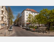 Eine städtische Straßenszene mit gepflasterter Fahrbahn, flankiert von mehrstöckigen Wohnhäusern mit hellen Fassaden. Im Vordergrund stehen Autos geparkt, während sich rechts ein kleiner Platz mit Bäumen und Grünflächen befindet.