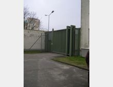 Großes grünes Metallschiebetor neben einer grauen Betonmauer und einem Maschendrahtzaun mit Stacheldraht, davor eine asphaltierte Zufahrt. Im Hintergrund stehen ein hoher Straßenmast, kahle Bäume und ein mehrstöckiges Gebäude.