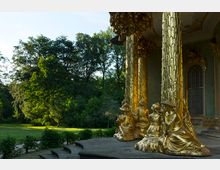 Goldene, detailreich verzierte Säulen mit sitzenden vergoldeten Skulpturen vor einem historischen Pavillon; im Hintergrund eine grüne Parklandschaft mit dichtem Baumbestand.