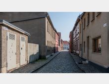 Schmale, gepflasterte Seitenstraße mit einfachen Wohngebäuden aus Putz, Fachwerk und Backstein. Die Straße ist von schlichten Häuserwänden ohne viel Verzierung gesäumt, mit vereinzelten Fenstern und Haustüren; im Hintergrund ein weiteres Gebäude mit roten Ziegeldächern.